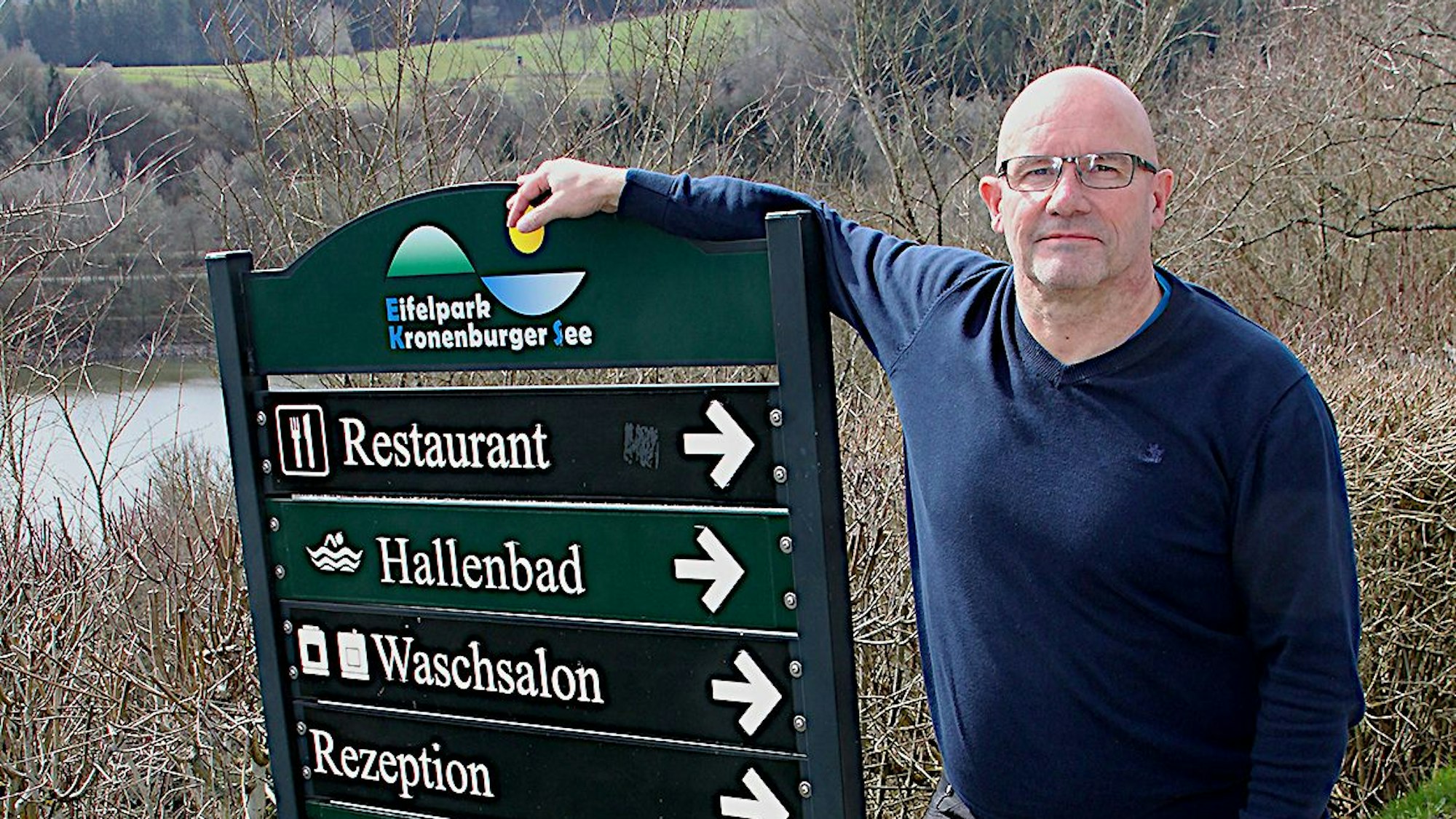 Das Bild zeigt einen Mann, der sich auf eine Hinweistafel mit Wegweisern des Ferienparks stützt.