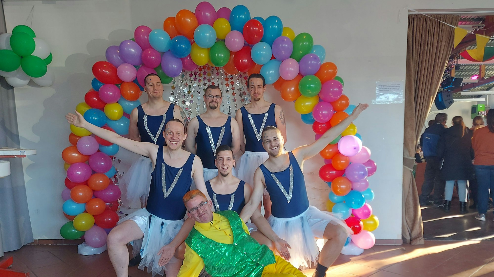 Sieben Männer in Kostümen posieren vor einem Halbkreis aus bunten Luftballons.