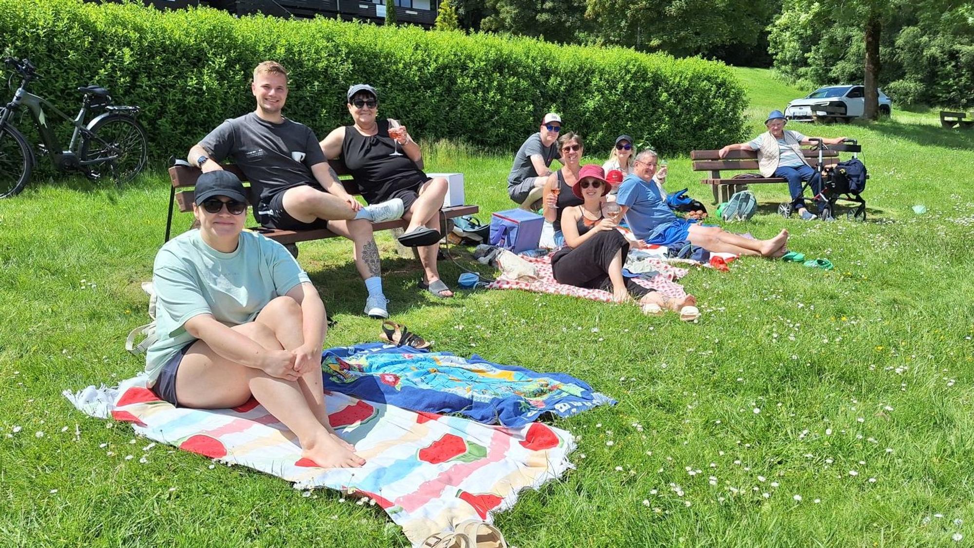 Das Bild zeigt Männer und Frauen, die sich auf einer Rasenfläche auf Sitzbänken und Decken niedergelassen haben.
