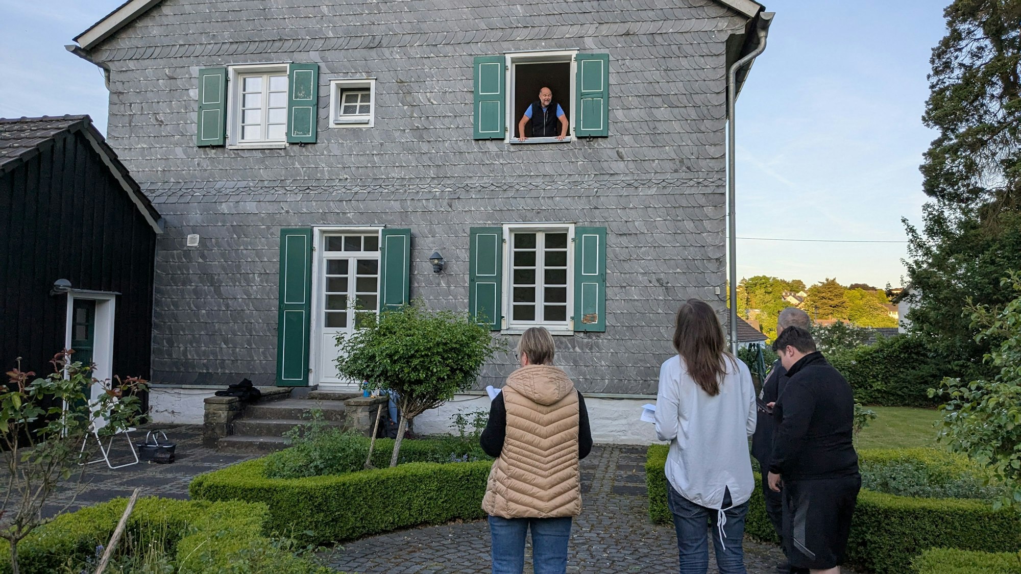 Drei Personen stehen vor einem geschieferten Gebäude, aus einem Fenster schaut ein Mann heraus.