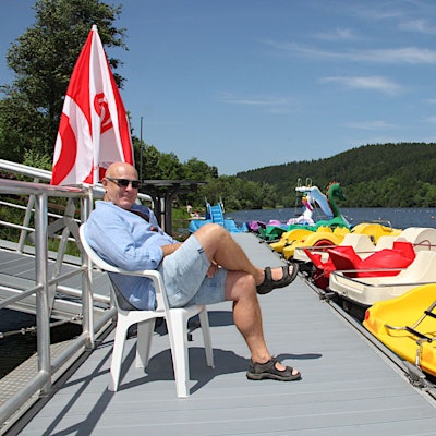Das Bild zeigt einen Mann in Shorts auf einem Steg, an dem Tretboote in verschiedenen Farben liegen.