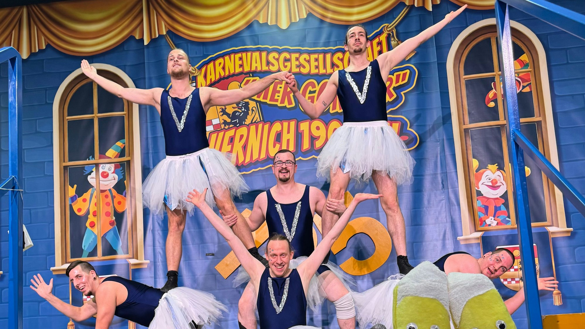 Sechs Männer in Tutus präsentieren auf einer Bühne eine Figur. Ihre Körper bilden mehrere Etagen.
