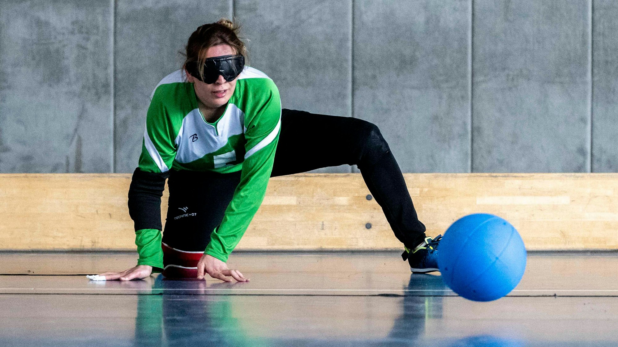 Eine Sportlerin mit dunkler Brille kniet auf dem Boden einer Turnhalle, ein blauer Ball rollt auf sie zu.