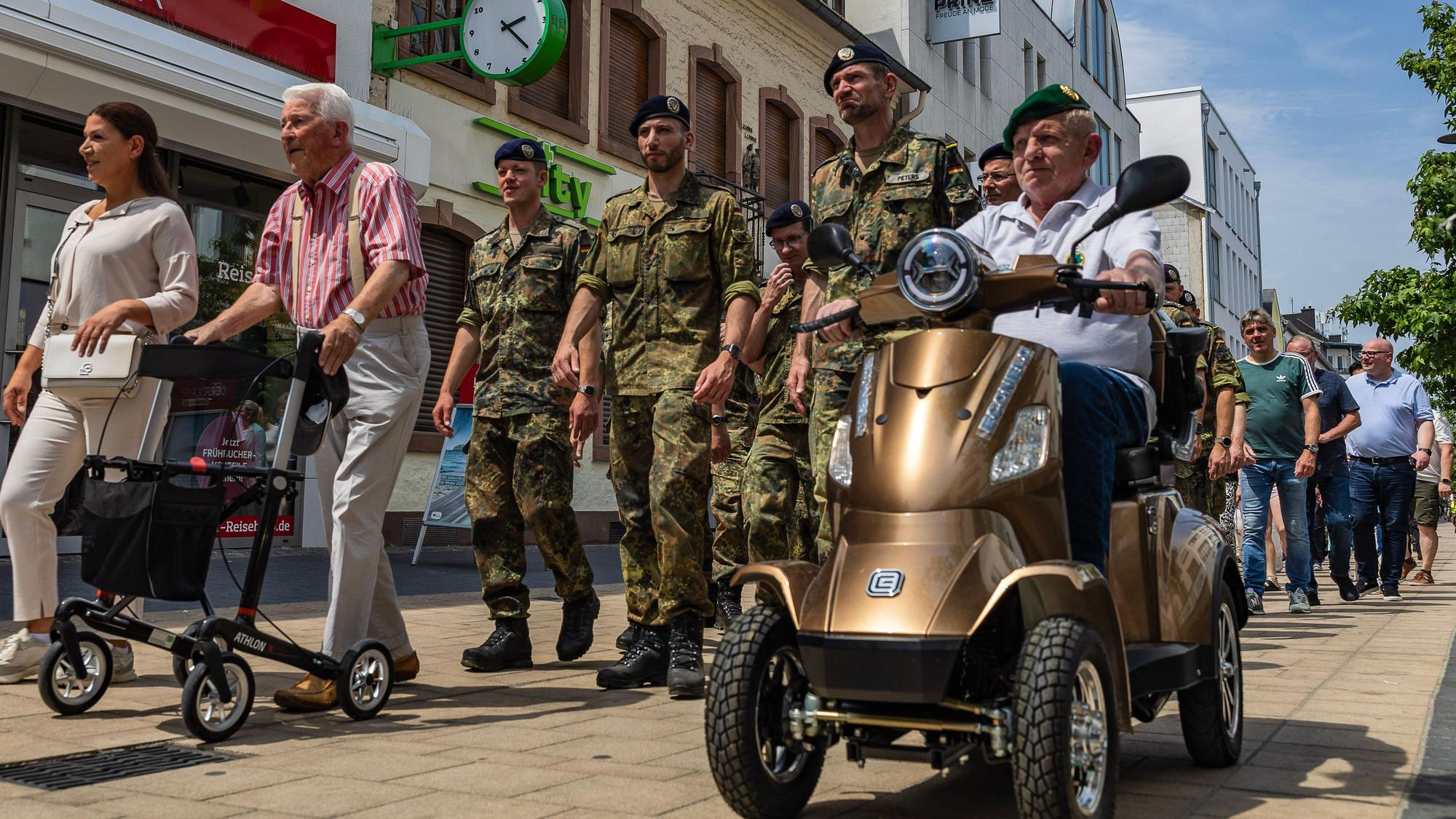 Soldaten in Feldanzügen marschieren auf der Neustraße. Vor ihnen ein Ex-Soldat mit Rollator und ein weiterer mit grünem Barett auf einem Elektromobil.