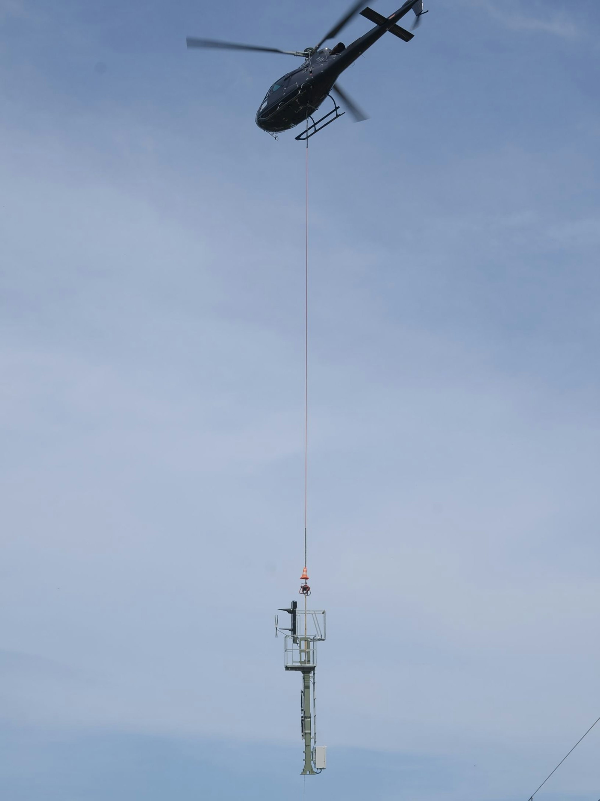 Ein Helikopter transportiert einen Mast an einem langen Stahlseil.