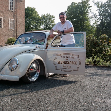 Das Foto zeigt einen Mann, der hinter der offenen Tür eines VW Käfers an seinem Wagen lehnt.