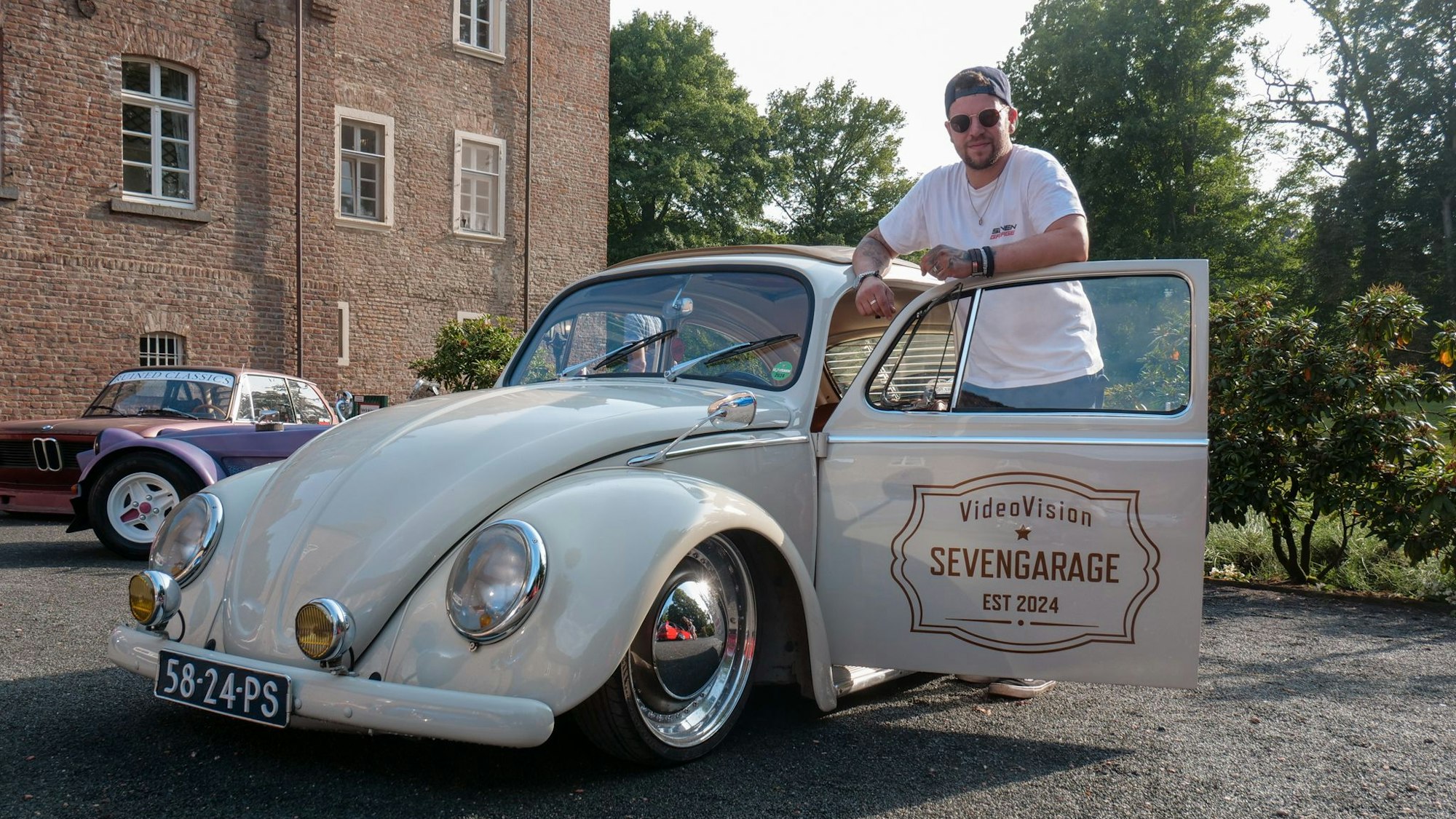 Das Foto zeigt einen Mann, der hinter der offenen Tür eines VW Käfers an seinem Wagen lehnt.