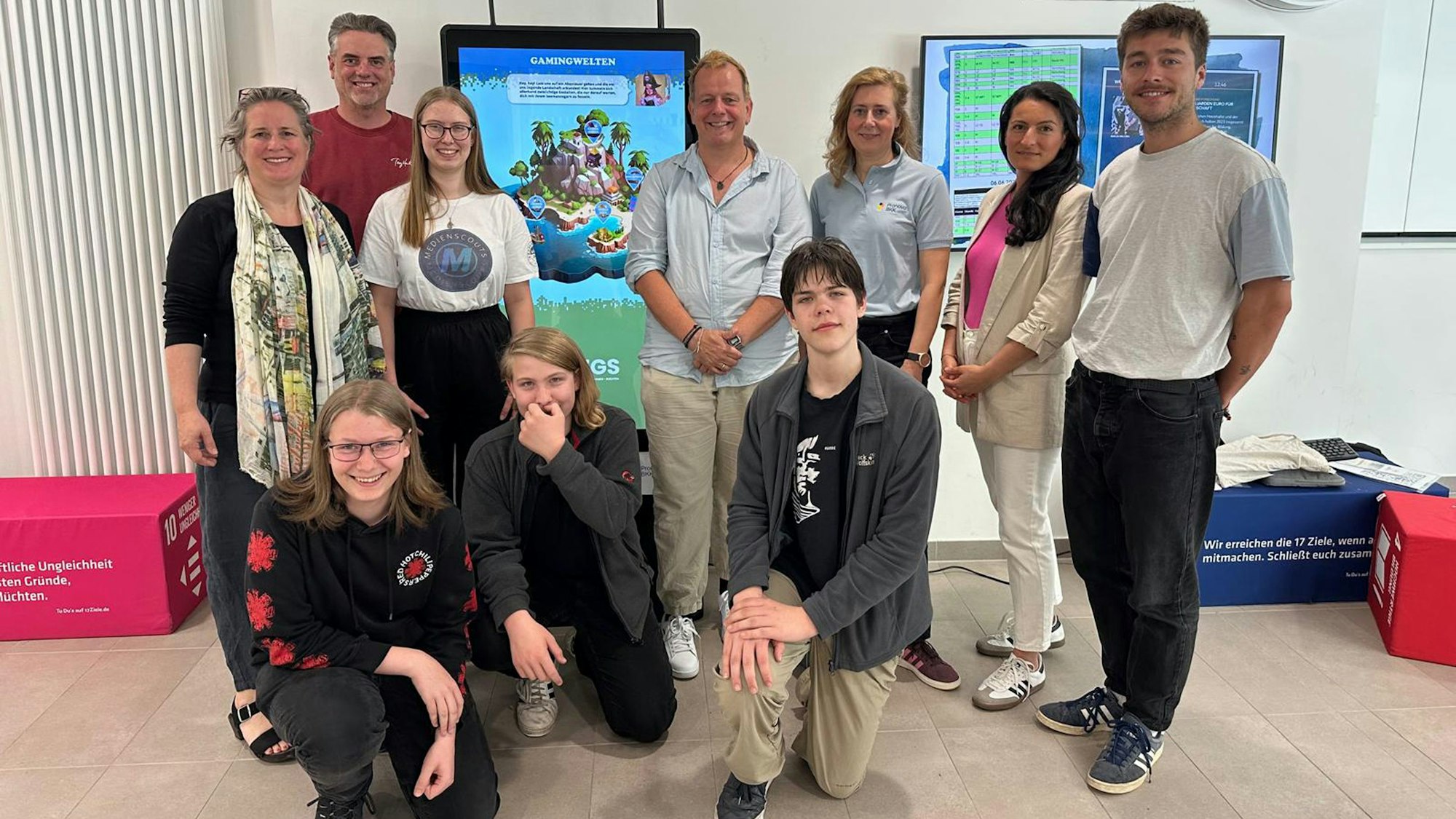 Eine Gruppe Menschen steht vor Monitoren, die an der Wand hängen, drei Schüler knien vor den sieben Stehenden.