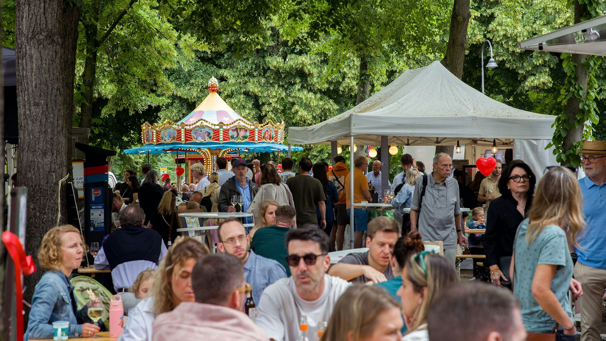 Menschen sitzen an Biertischen, im Hintergrund ein Kinderkarussell.