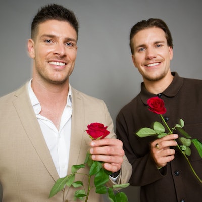 Die neuen „RTL-Bachelors“ Martin Braun (l.) und Felix Stein (r.) halten Rosen in der Hand und lächeln vor einer Wand stehend in Richtung Kamera.