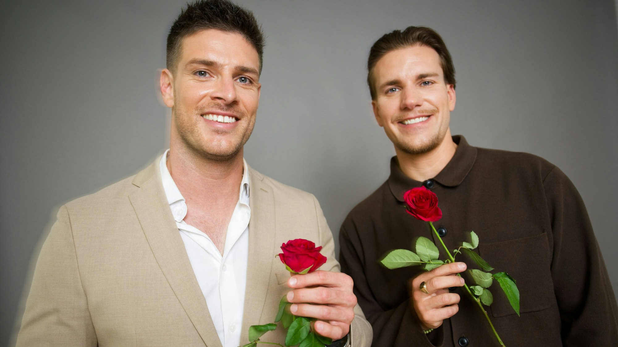Die neuen „RTL-Bachelors“ Martin Braun (l.) und Felix Stein (r.) halten Rosen in der Hand und lächeln vor einer Wand stehend in Richtung Kamera.