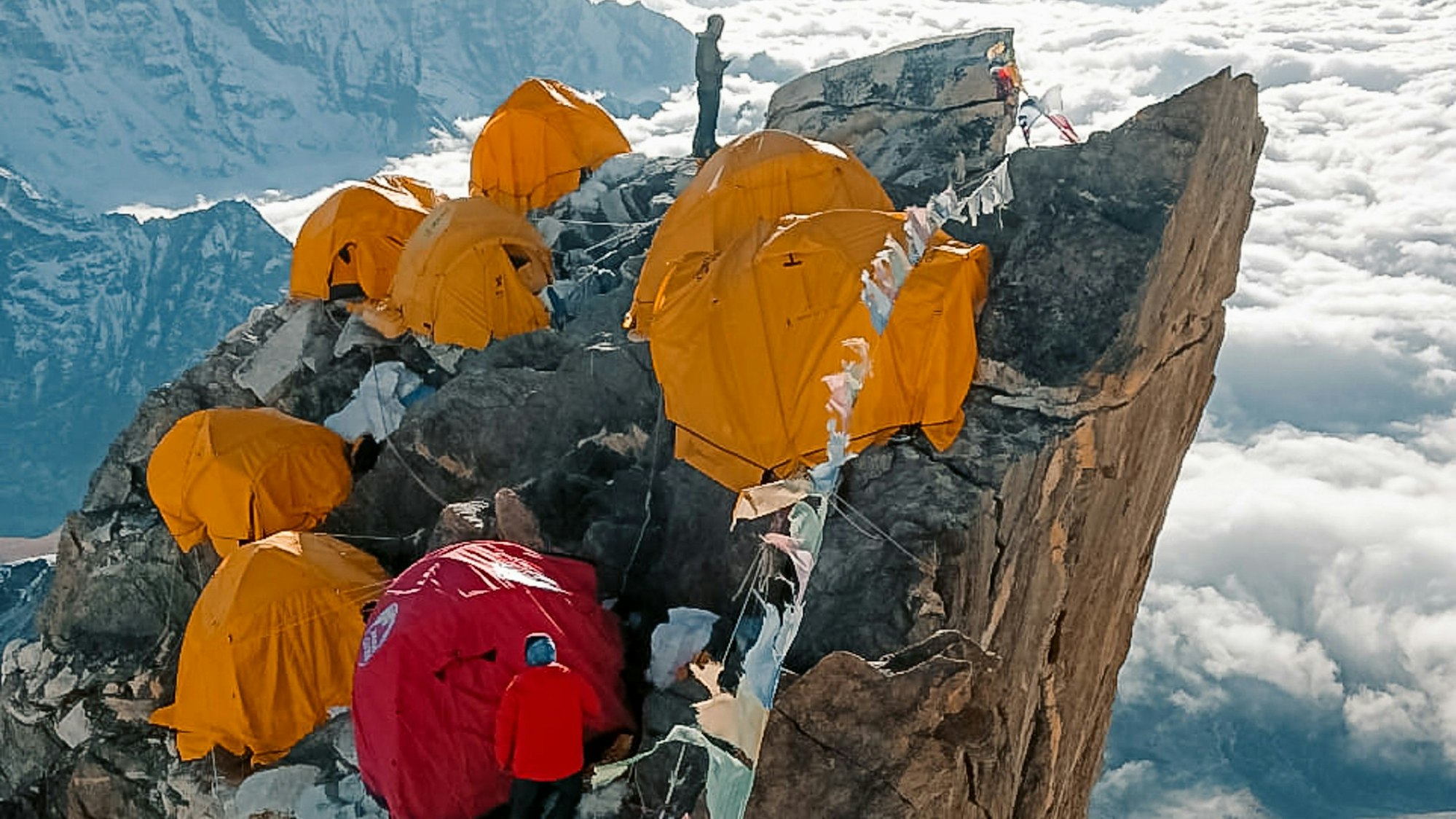 Auf einem Bergvorsprung stehen mehrere Zelte, dazwischen zwei Personen.