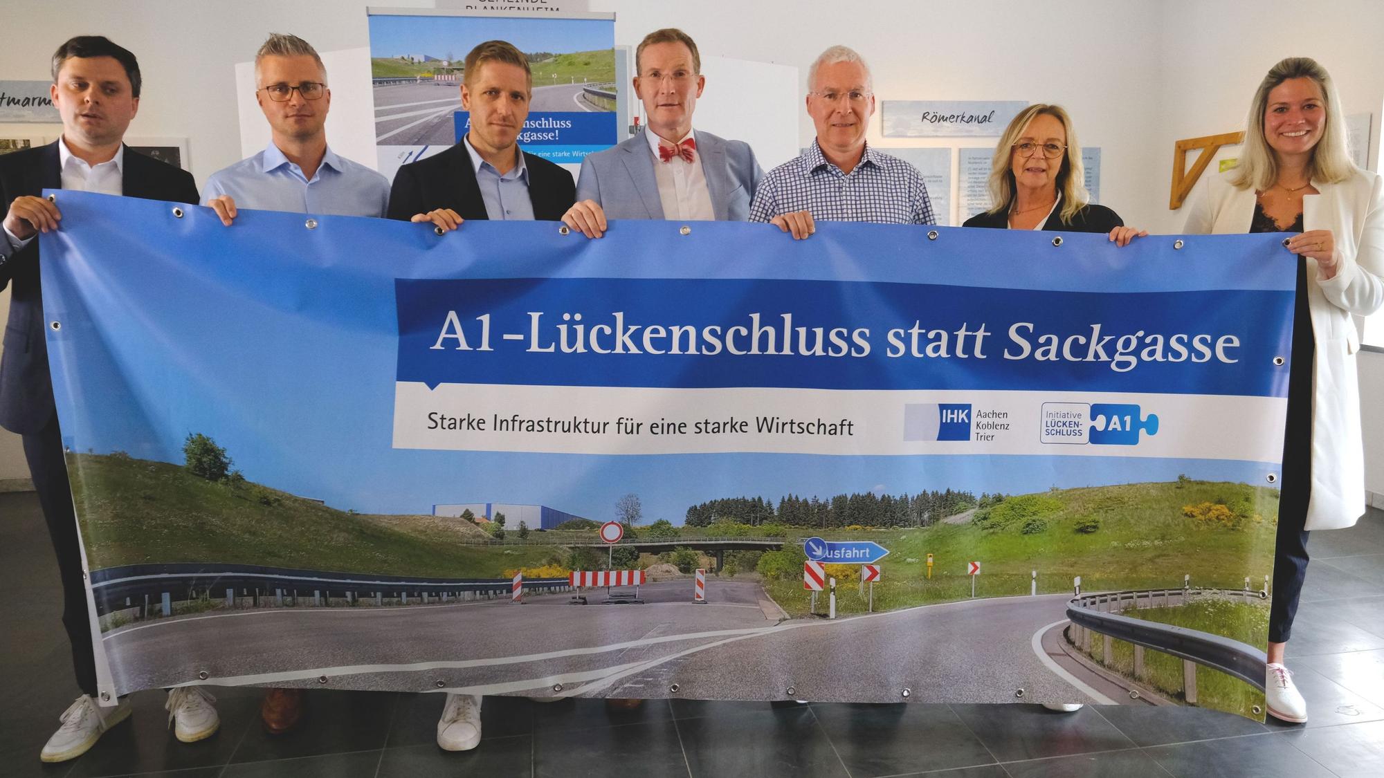 IHK-Aktion zum A1-Lückenschluss, ein entsprechendes Transparent halten v.l.n.r.): Philipp Piecha (IHK Aachen), Daniel Claßen (Spedition Berners), Markus Ramers, Michael F. Bayer (IHK Aachen), Stephan Mahlert (Pappen Olef GmbH), Sabine Preiser-Marian (Bürgermeisterin Bad Münstereifel), Jennifer Meuren (Bürgermeisterin Blankenheim).