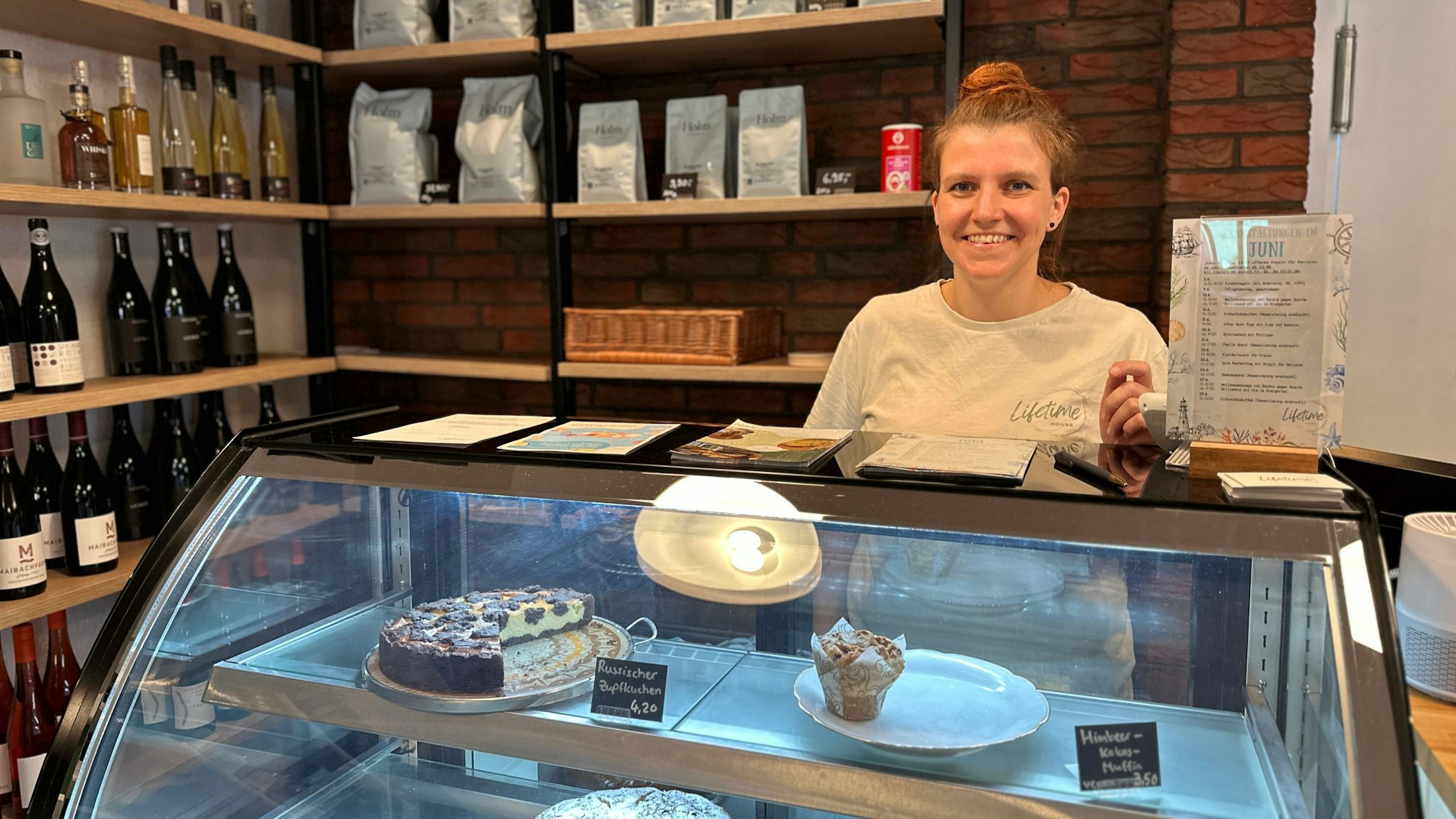 Ein Frau steht hinter ein Glas-Theke, die mit Kuchen bestückt ist. Im Regal hinter ihr stehen Weinflaschen und Packungen mit Kaffeepulver.