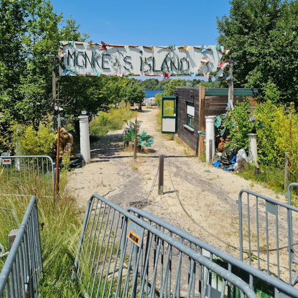 Der verlassene Eingang zum Beachclub „Monkey's Island“. An einem Häuschen steht die Tür auf, ein Rollo ist halb heruntergelassen. Pflanzen wachsen wild links und rechts des Weges, Absperrgitter stehen kreuz und quer auf dem Weg. Auf dem großen Schild mit der Aufschrift „Monkey's Island“ fehlt das y.