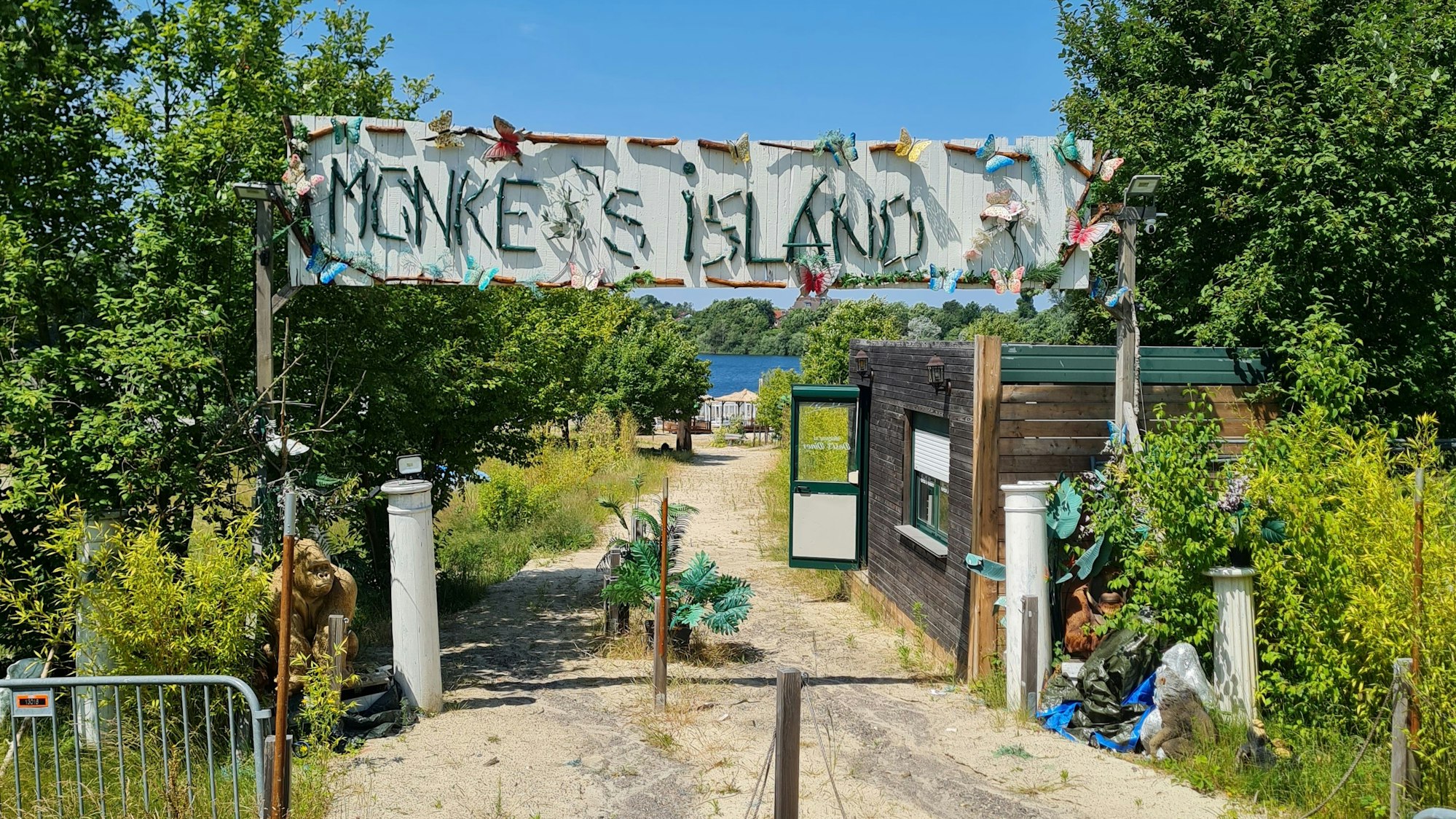 Der verlassene Eingang zum Beachclub „Monkey's Island“. An einem Häuschen steht die Tür auf, ein Rollo ist halb heruntergelassen. Pflanzen wachsen wild links und rechts des Weges, Absperrgitter stehen kreuz und quer auf dem Weg. Auf dem großen Schild mit der Aufschrift „Monkey's Island“ fehlt das y.