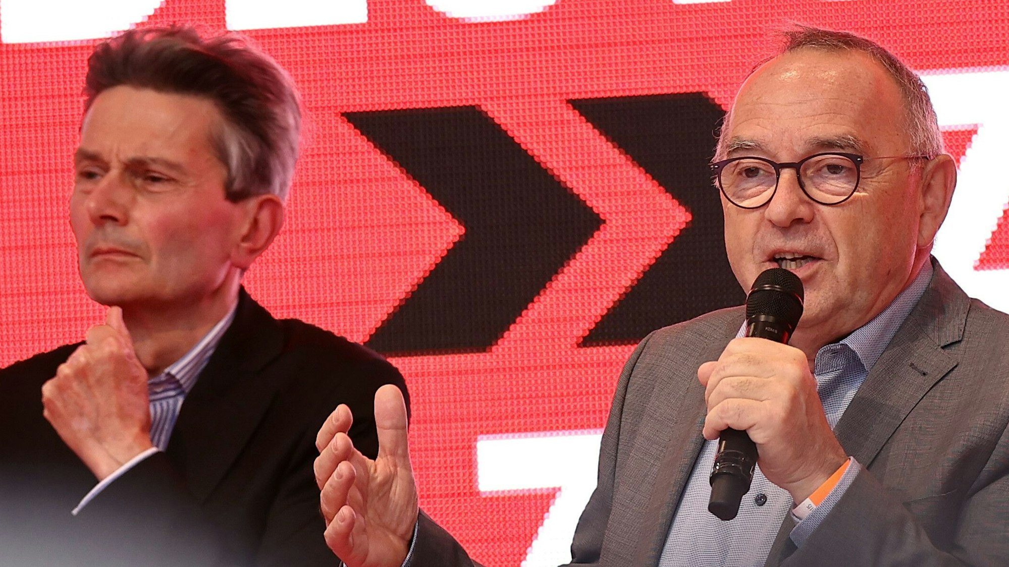 Rolf Mützenich (l.) und Norbert Walter-Borjans stehen vor einem rotgründigen Hintergrund. Walter-Borjans hat ein Mikrofon in der Hand und spricht, Mützenich hört zu.