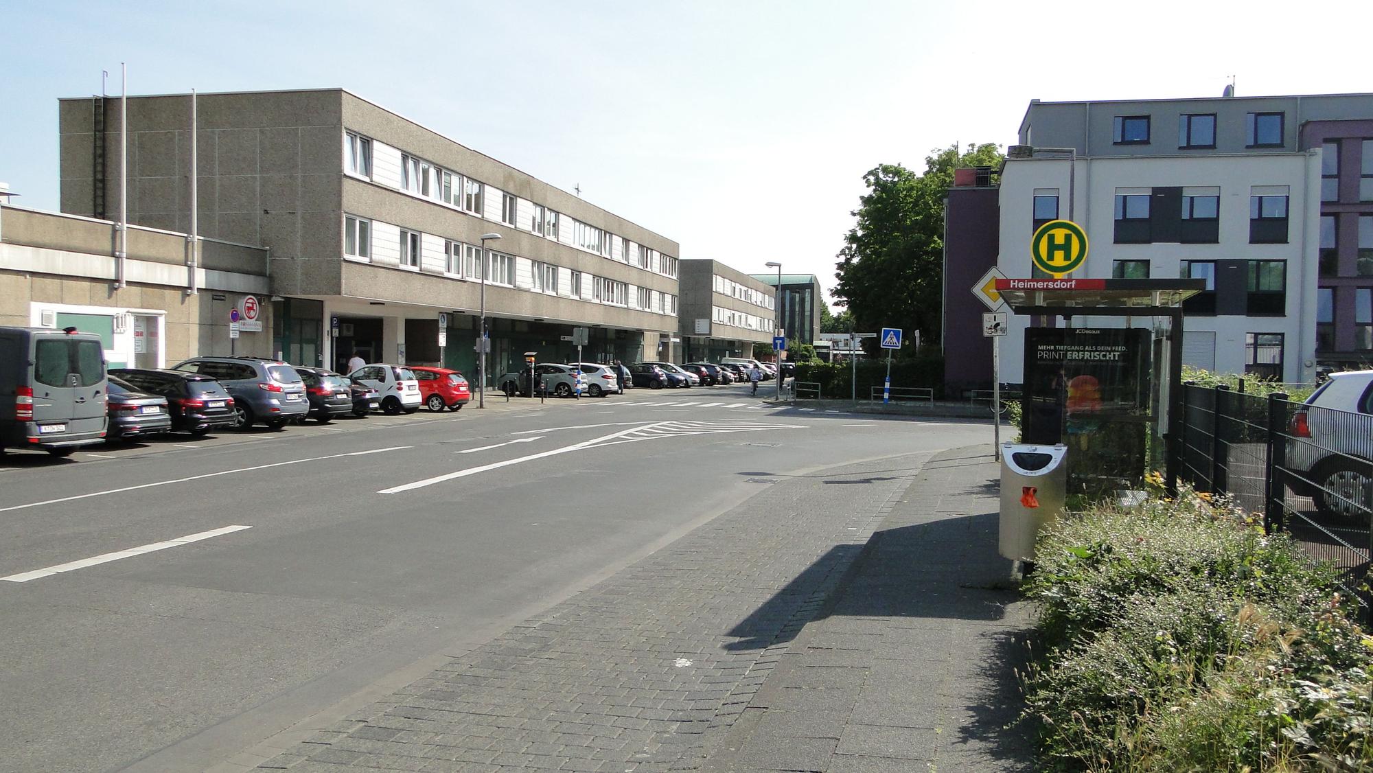 An einer Straße liegt eine Bushaltestelle, auf der Straßenseite gegenüber ist ein schmuckloser dreistöckiger Bau, der Haselnusshof, zu sehen.