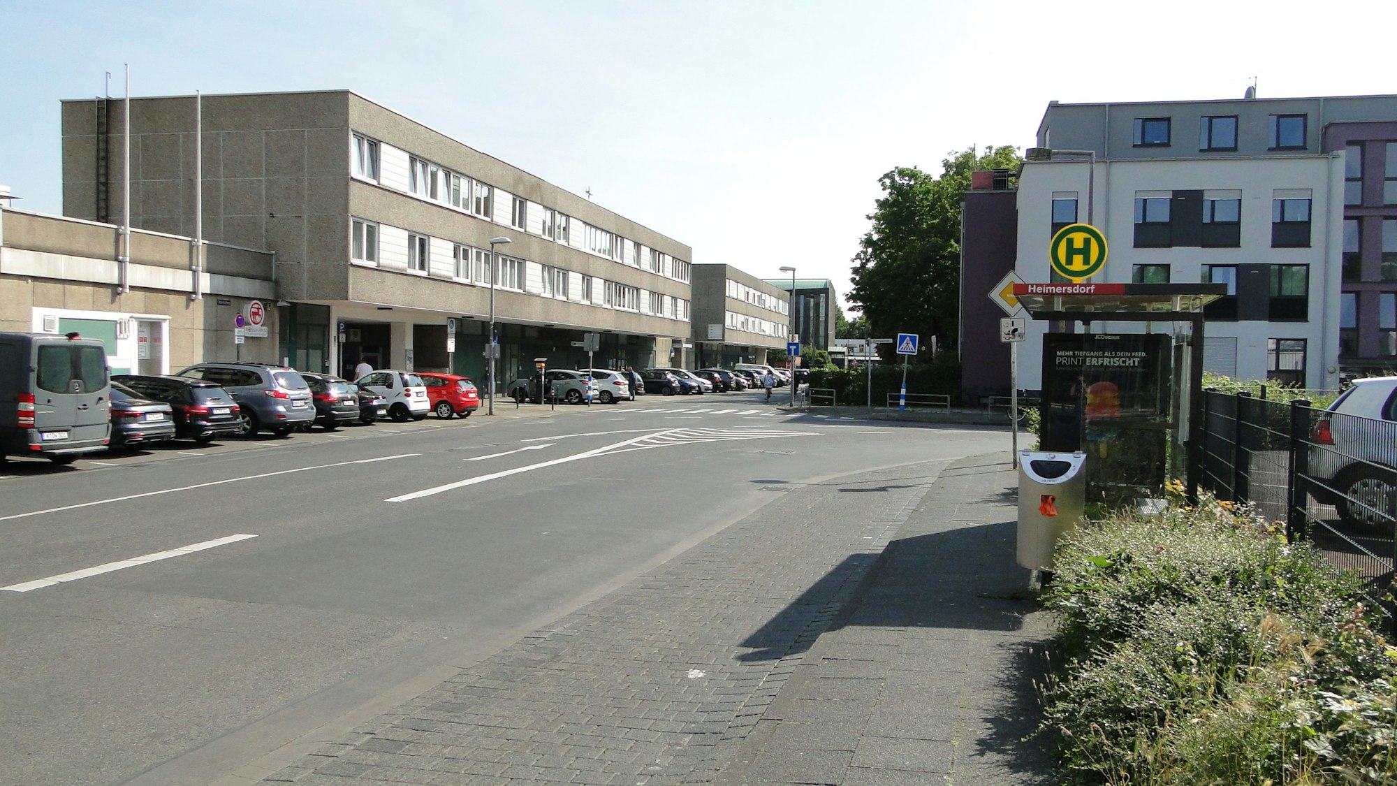 An einer Straße liegt eine Bushaltestelle, auf der Straßenseite gegenüber ist ein schmuckloser dreistöckiger Bau, der Haselnusshof, zu sehen.