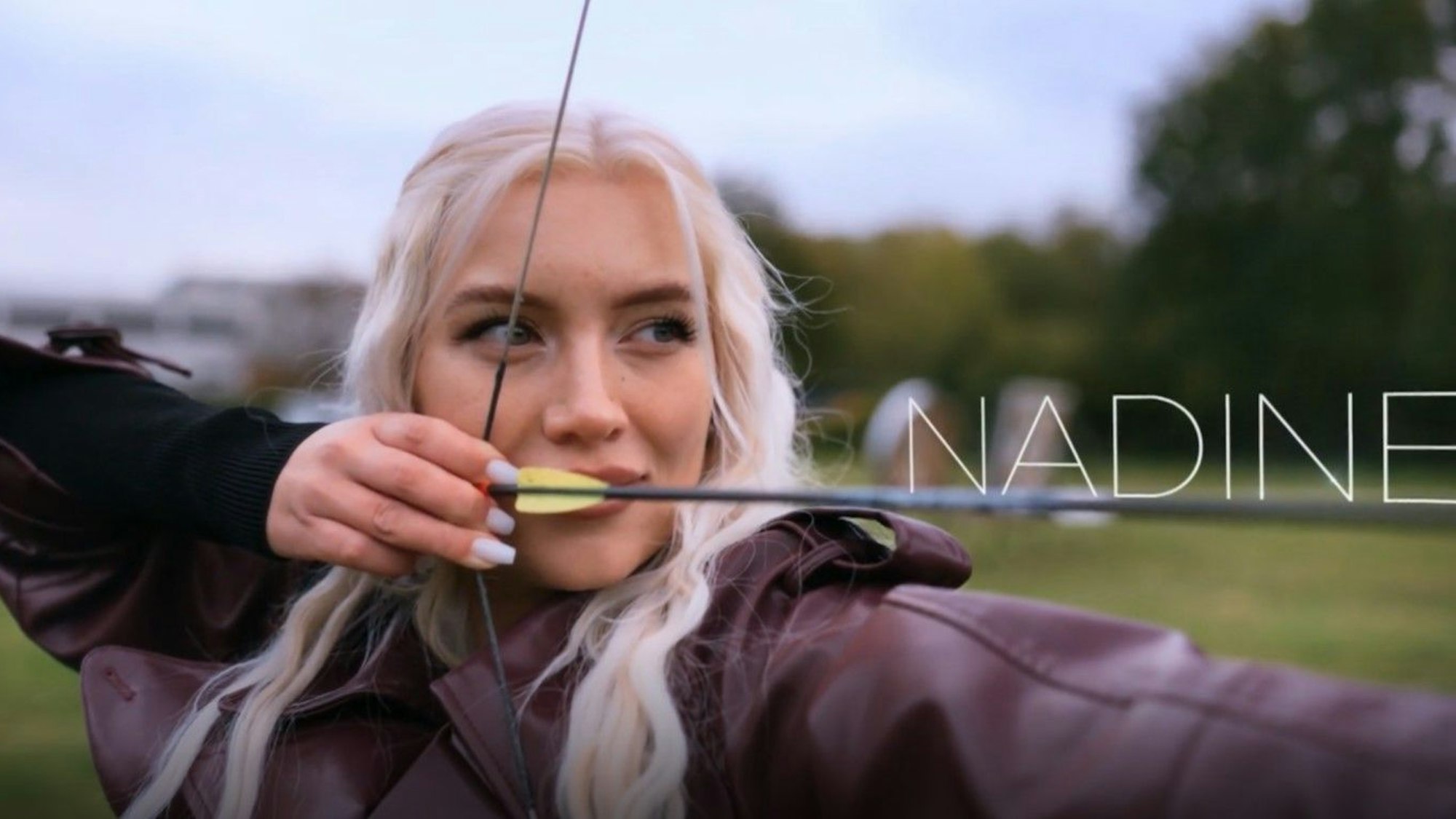 Kandidatin Nadine spannt einen Pfeil in einen Bogen und ist kurz davor, den Pfeil auf eine Scheibe abzufeuern. Close Up Einstellung der Kamera, Namensschriftzug „Nadine“ mittig rechts.
