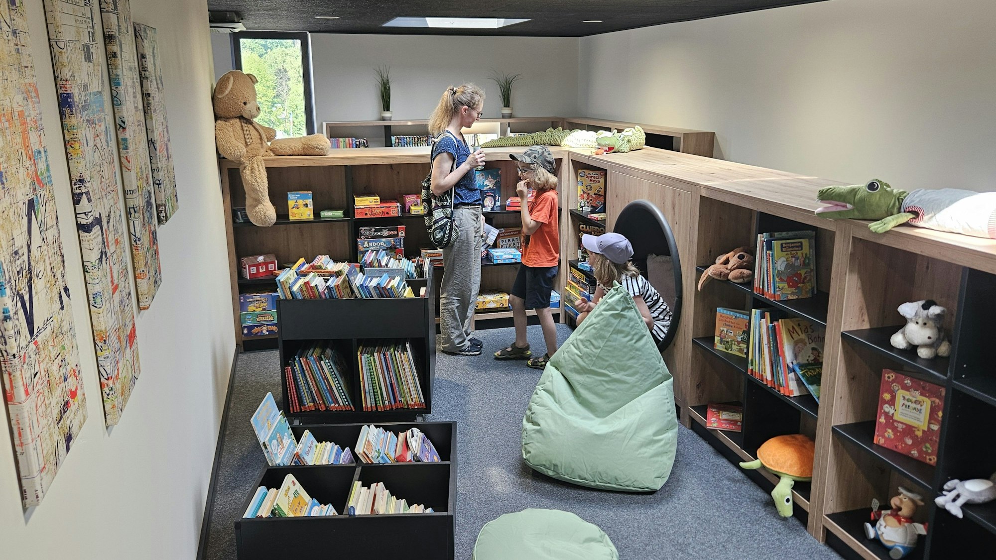 Blick auf die Spieleecke mit Büchern und Plüschfiguren. Zwei Kinder und eine Frau schauen sich die Angebote an.