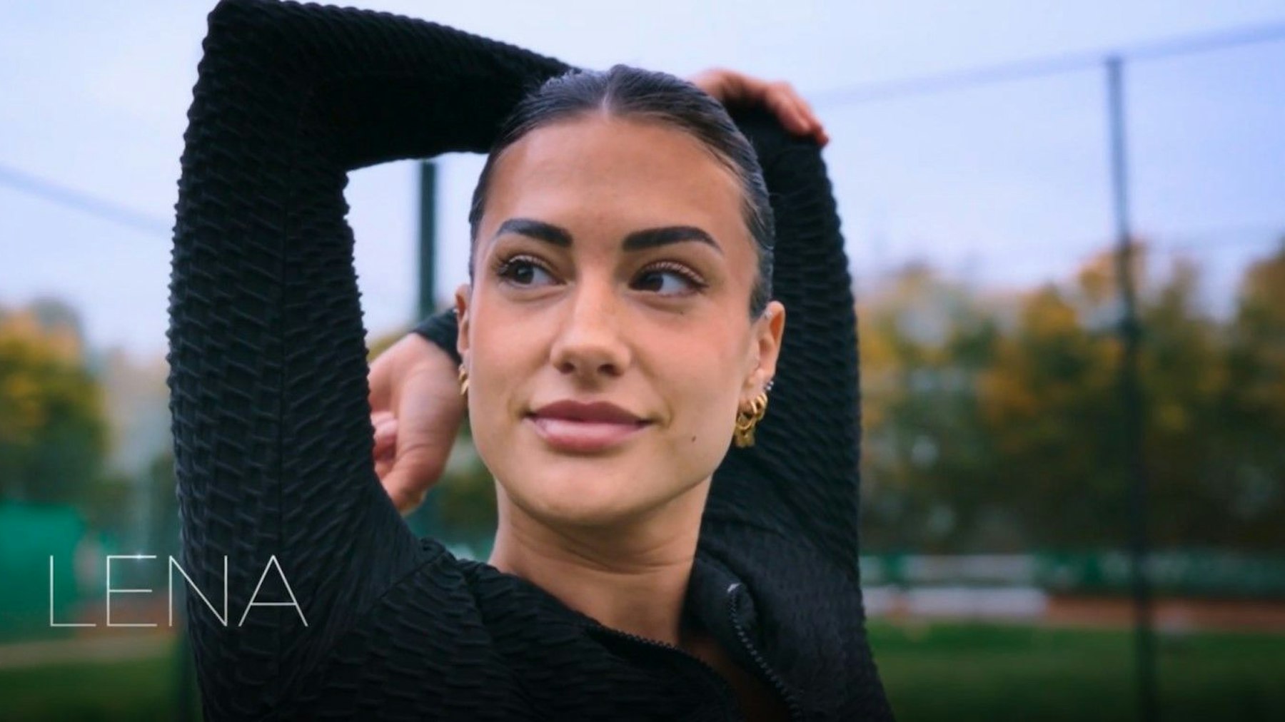 „Bachelor“-Kandidatin Lena (Namensschriftzug unten links) dehnt sich vor einem Tennisplatz. Close Up Einstellung.