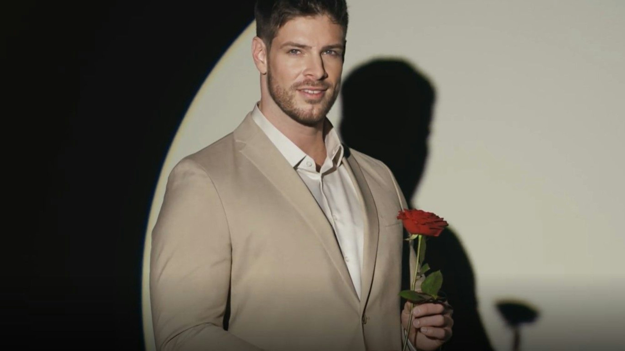 Einer der beiden Bachelor, Martin Braun, schaut in Richtung der Kamera. Er trägt einen hellbraunen Anzug und hält eine rote Rose in der linken Hand.