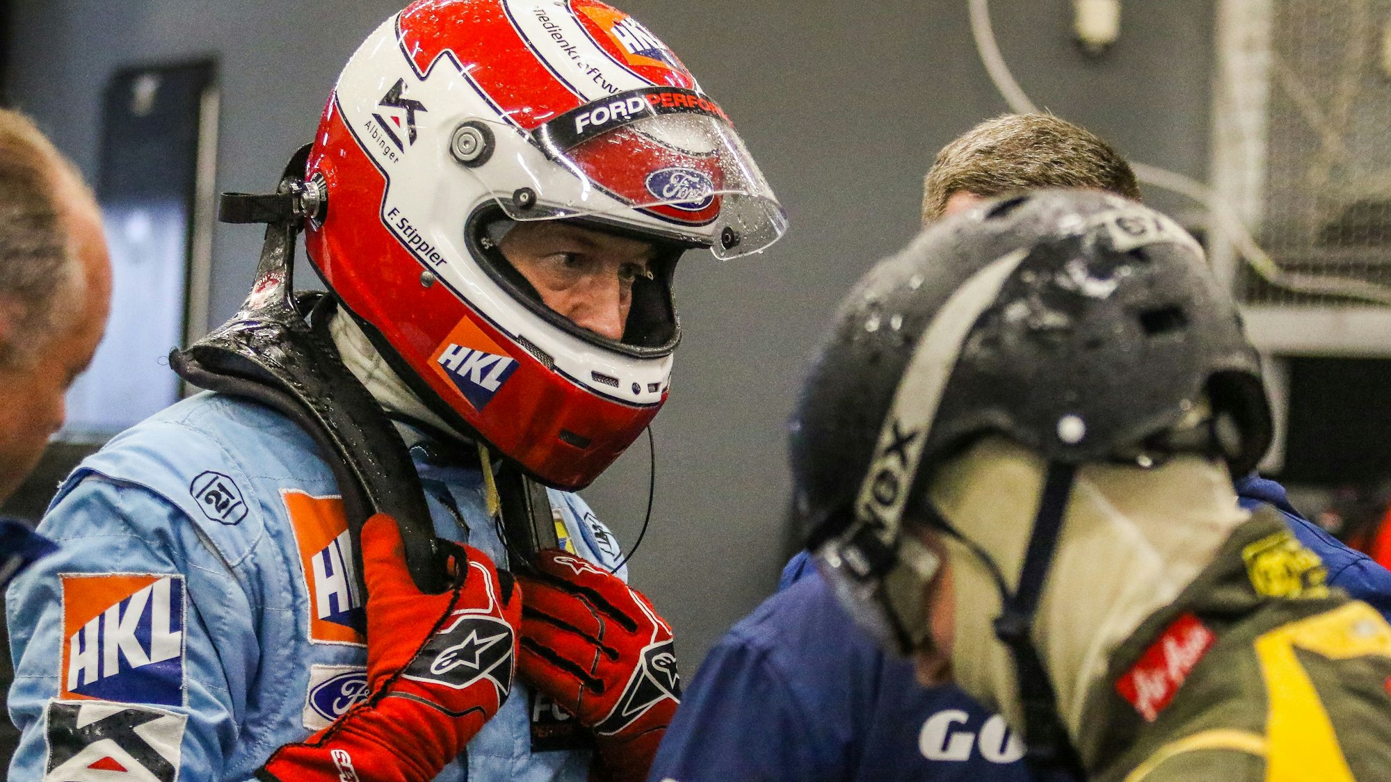 Frank Stippler im hellblauen Overall trägt einen rot-weißen Helm. Seine Hände stecken in roten Handschuhen. Er spricht in der Box mit einem Techniker.
