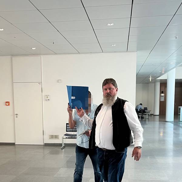Ein Mann mit Bart schaut in die Kamera. Ein zweiter Mann, Angeklagter in einem Prozess, hält sich einen blauen Aktenordner vors Gesicht.