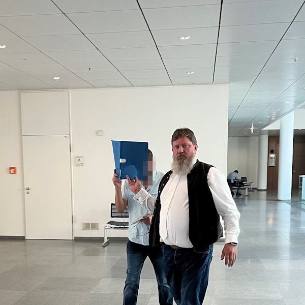 Ein Mann mit Bart schaut in die Kamera. Ein zweiter Mann, Angeklagter in einem Prozess, hält sich einen blauen Aktenordner vors Gesicht.