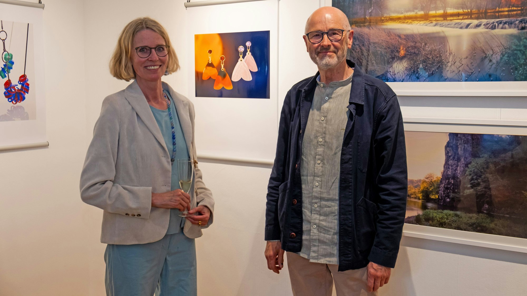 Eine Frau mit blondem Haar und ein Mann mit Glatze, Bart und Brille stehen vor einer Wand, an der Bilder hängen.