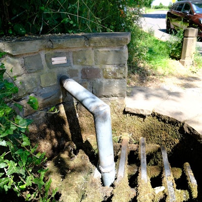 An einem Brunnen ist das Auslassrohr des Wassers bis unter ein Gitter verlängert worden, um die Wasserentnahme zu verhindern.