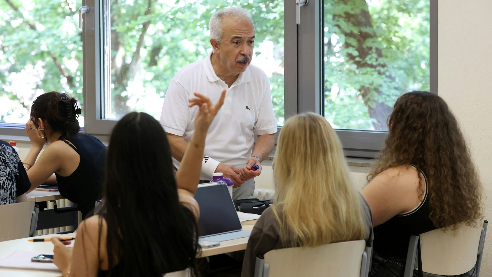 Eine Unterrichtsszene ist zu sehen, drei Mädchen sitzen in der Stuhlreihe, eine meldet sich.