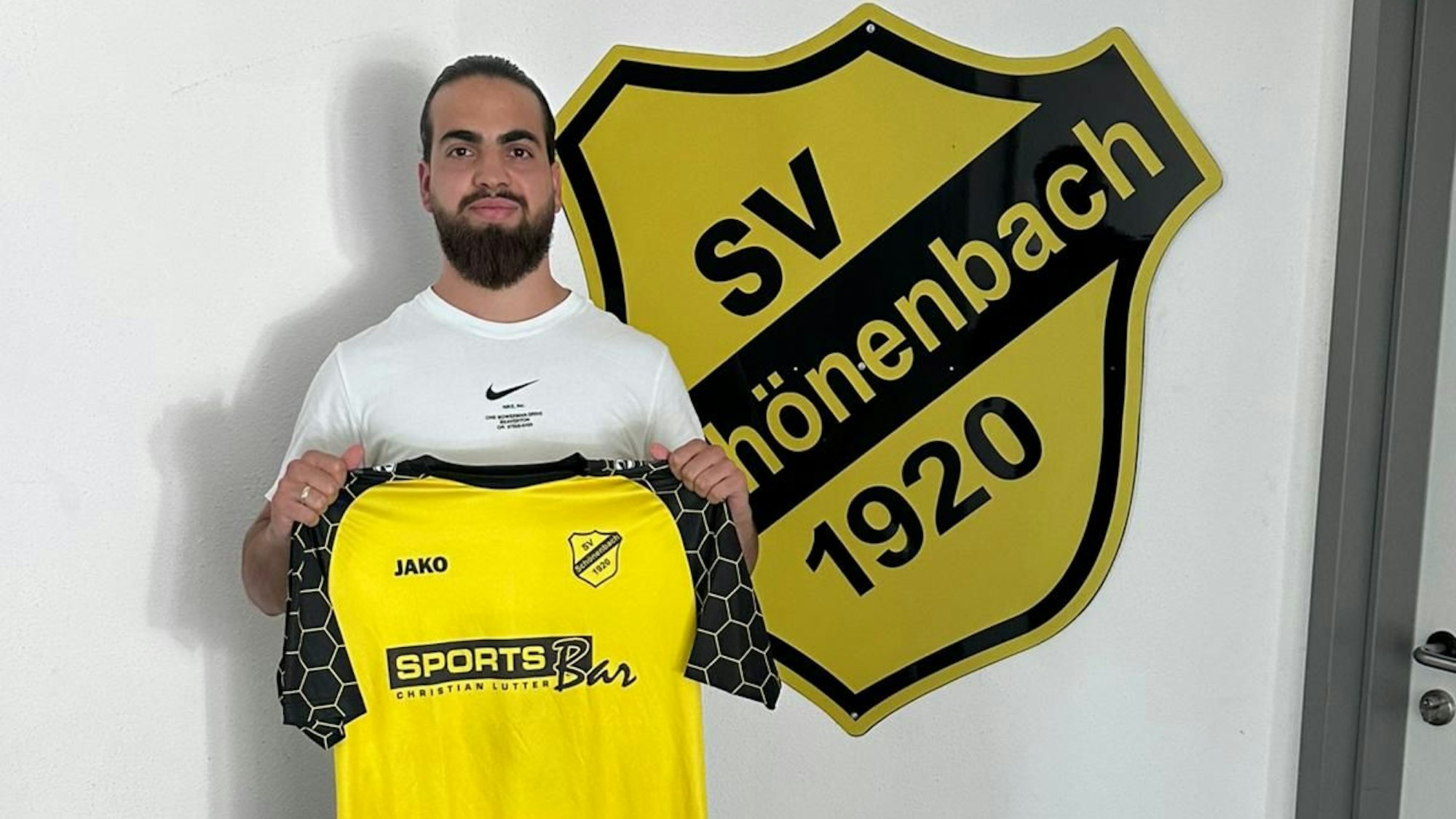 Louis Vedder steht mit dem Schönenbacher Trikot in der Hand vor dem Vereinsabzeichen.