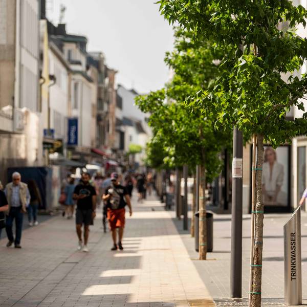 Der Blick in die Neustraße zeigt im Vordergrund einen Wasserspender, dahinter eine Baumreihe und Passantinnen und Passanten.