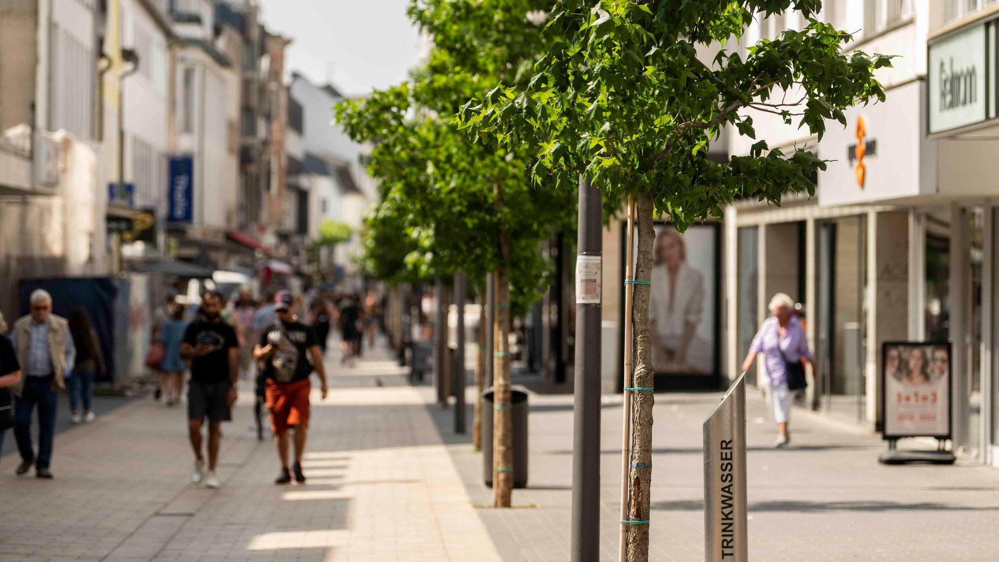 Der Blick in die Neustraße zeigt im Vordergrund einen Wasserspender, dahinter eine Baumreihe und Passantinnen und Passanten.
