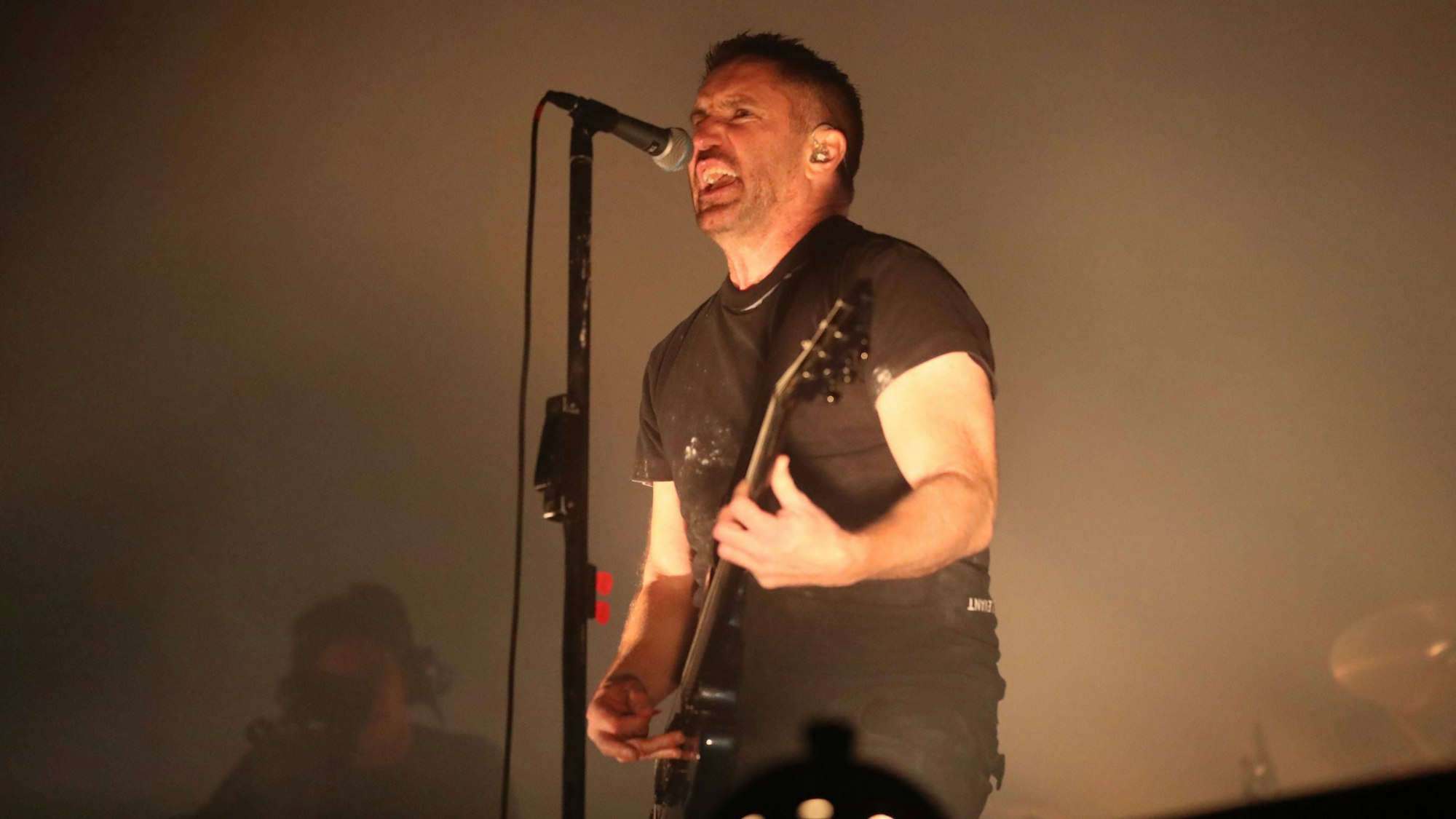 Trent Reznor steht Gitarre spielend vor einem Mikrofonständer und singt. Er trägt ein schwarzes T-Shirt und schwarze Jeans.