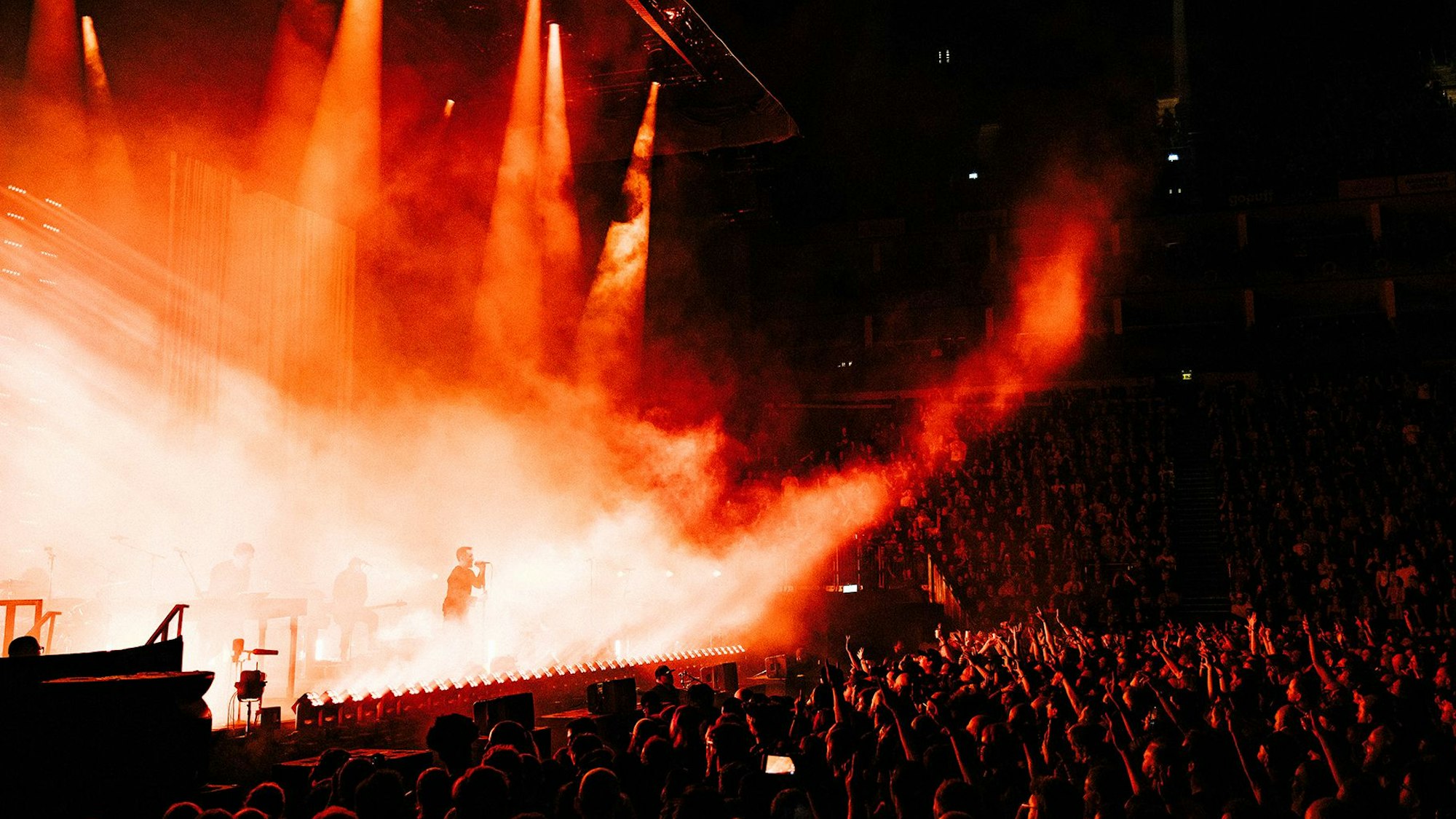 Trent Reznor hält einen Mikrofonständer und singt am Rand der in rotes Licht und Nebel getauchten Bühne. Vor ihm halten Fans die Arme hoch.