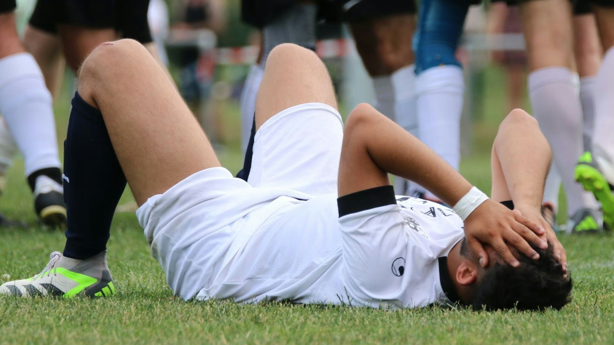 Ein Fußballer in weißer Spielkleidung liegt mit angezogenen Beinen auf dem Rücken und hält sich die Hände vors Gesicht.