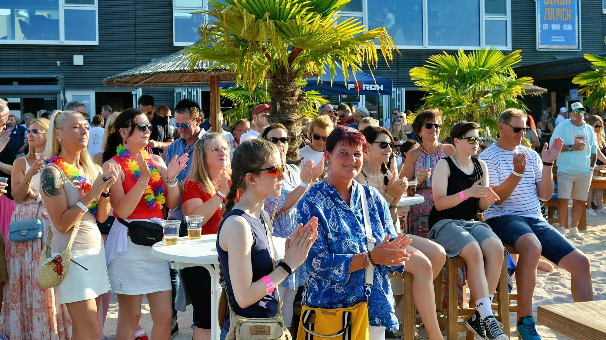 Das Bild zeigt Männer und Frauen in Sommerbekleidung im Freien. Viele von ihnen klatschen im Rhythmus der Musik.