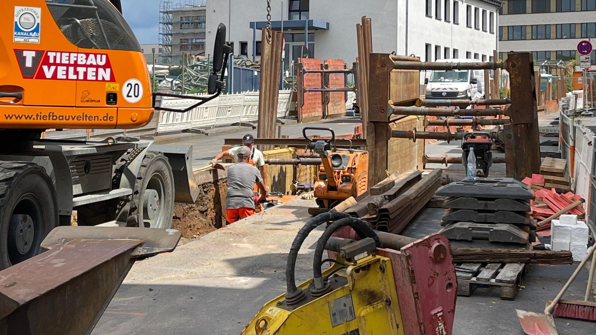 Das Bild zeigt eine Baustelle mit aufgebrochener Straße, Bagger und Arbeitern.