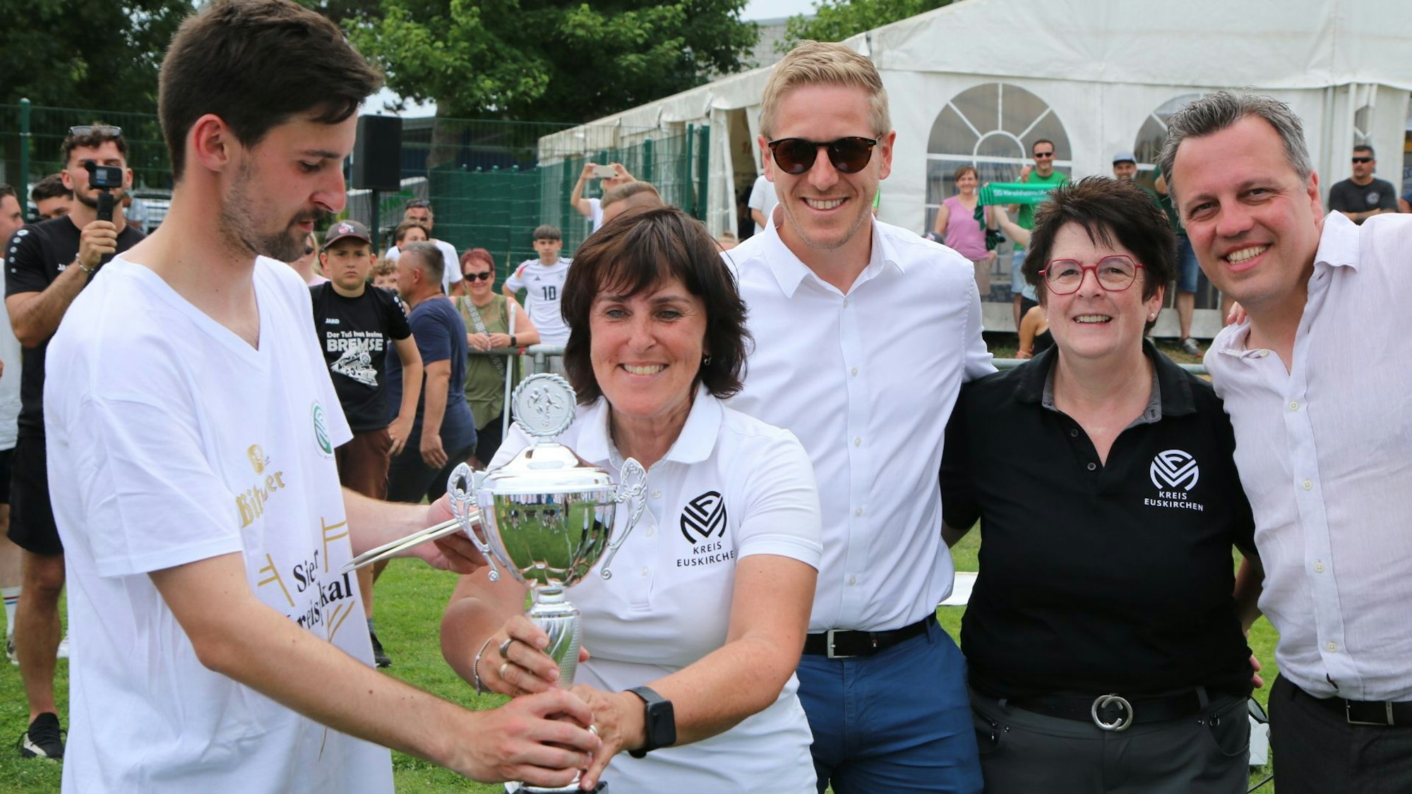 Fußballkreisvorsitzende Doris Mager überreicht ihrem Sohn, FlaKi-Kapitän Lukas Mager, den Pokal. Dahinter stehen Landrat Markus Ramers, Marion Knobbe vom Fußballkreis und Euskirchens Bürgermeister Sacha Reichelt.