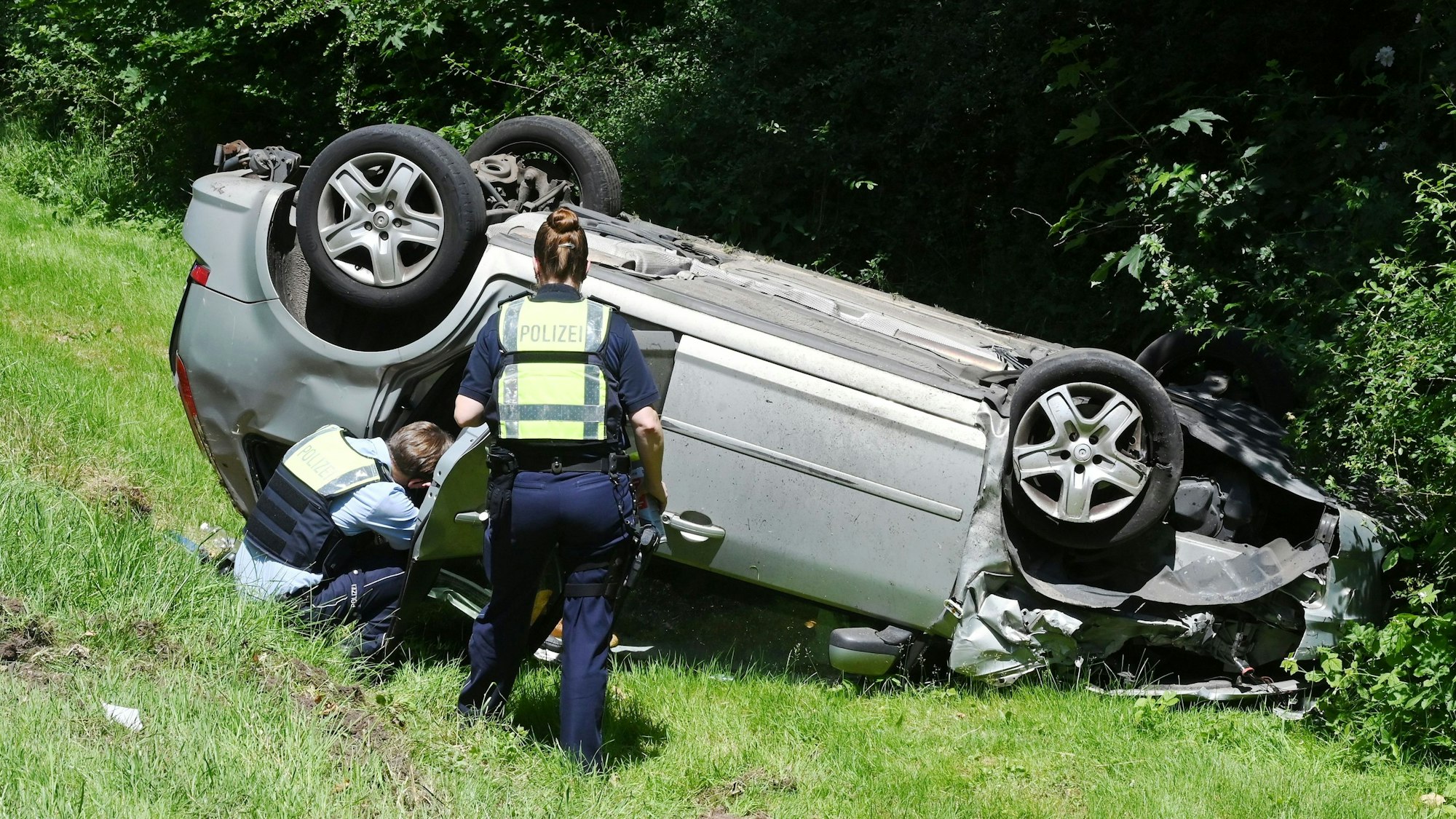 Das Bild zeigt das Auto, das auf dem Dach im Straßengraben liegen blieb. Eine Polizistin und ein Polizist untersuchen das Fahrzeug.