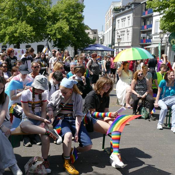 Menschen sitzen in Regenbogenfarben und mit Pride-Flaggen auf Bänken am Siegburger Markt.