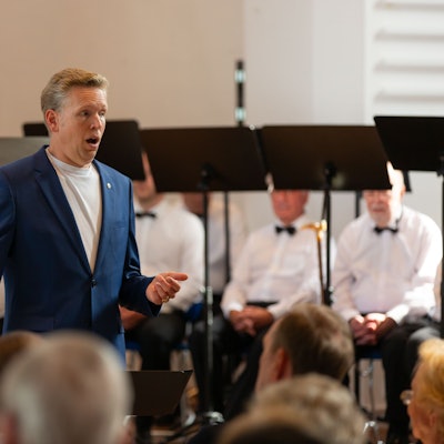 Ein Sänger steht am Flügel, dahinter sitzende Chorsänger.