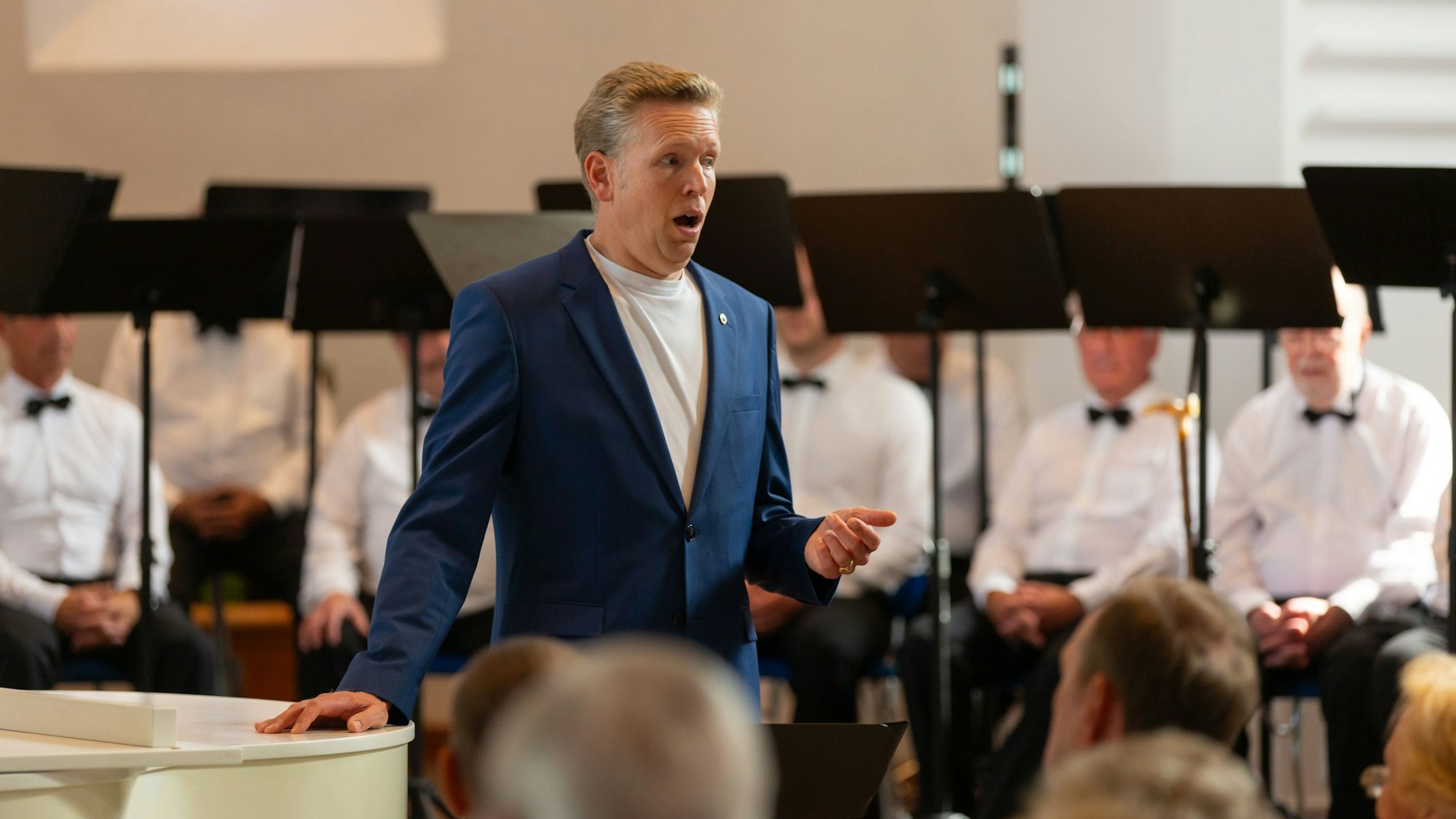 Ein Sänger steht am Flügel, dahinter sitzende Chorsänger.