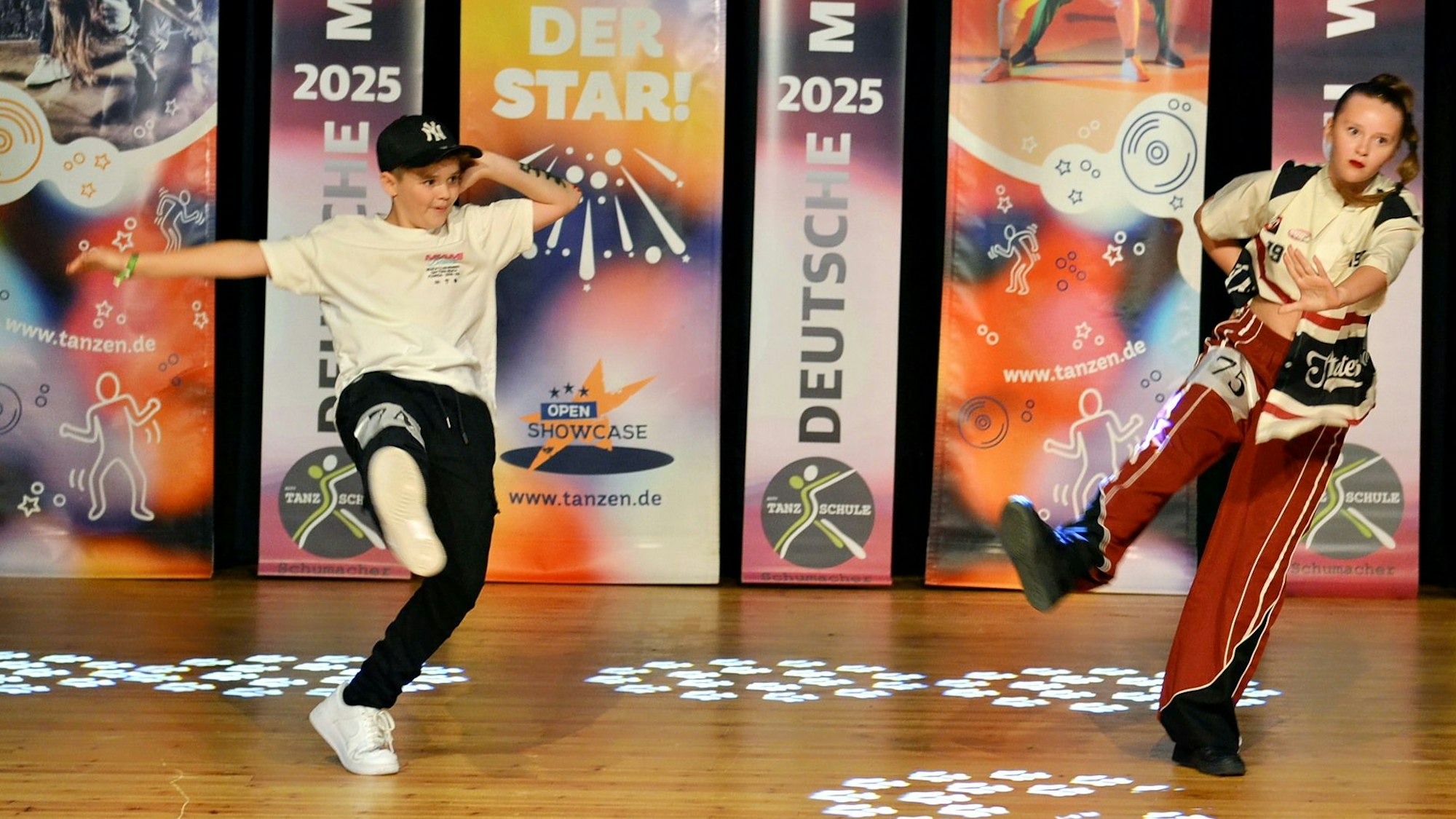 Zwei Jungs, einer mit Kappe und schwarzer Hose, einer mit roter Hose, zeigen Tanzfiguren.