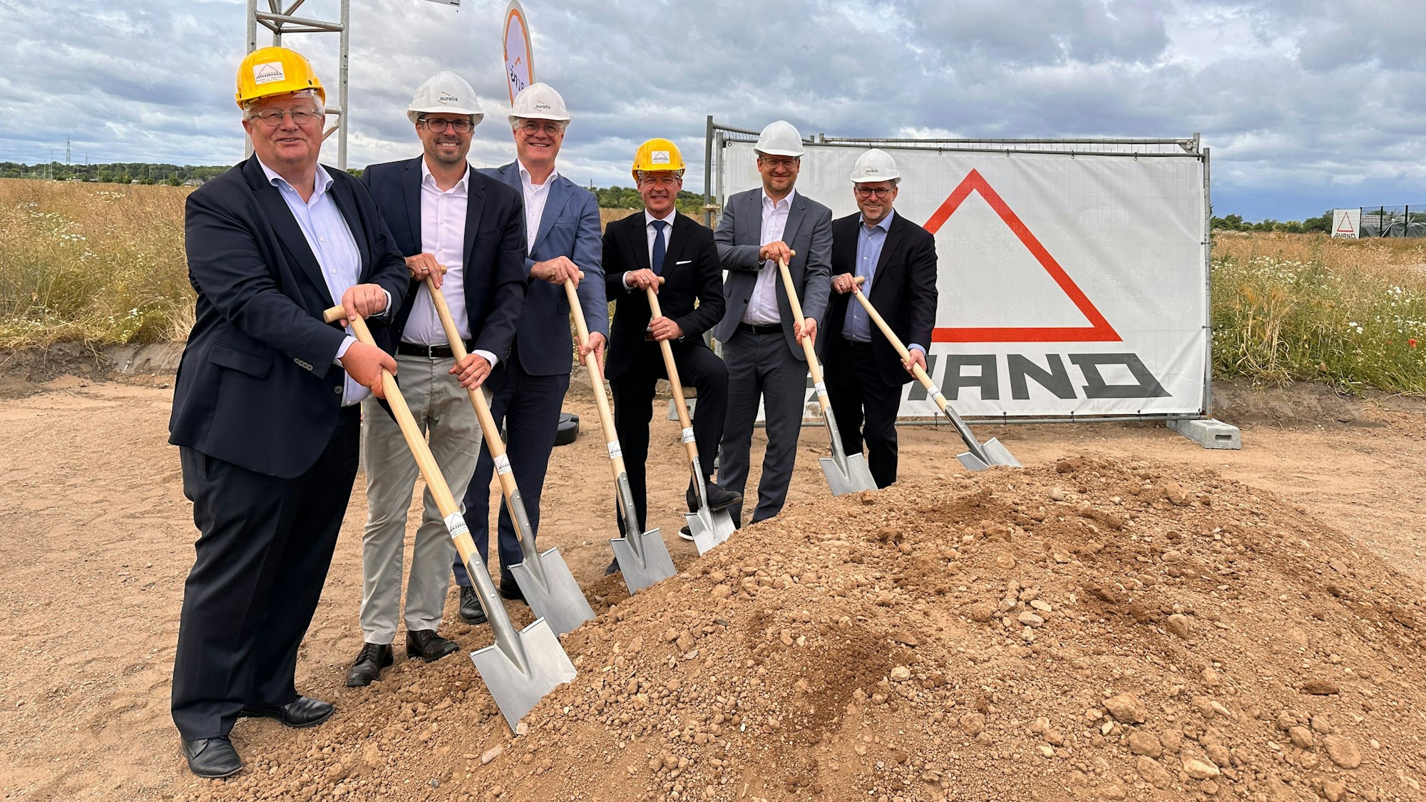 Sechs Männer mit Spaten stehen in einer Reihe neben einem aufgeschütteten Sandhügel.
