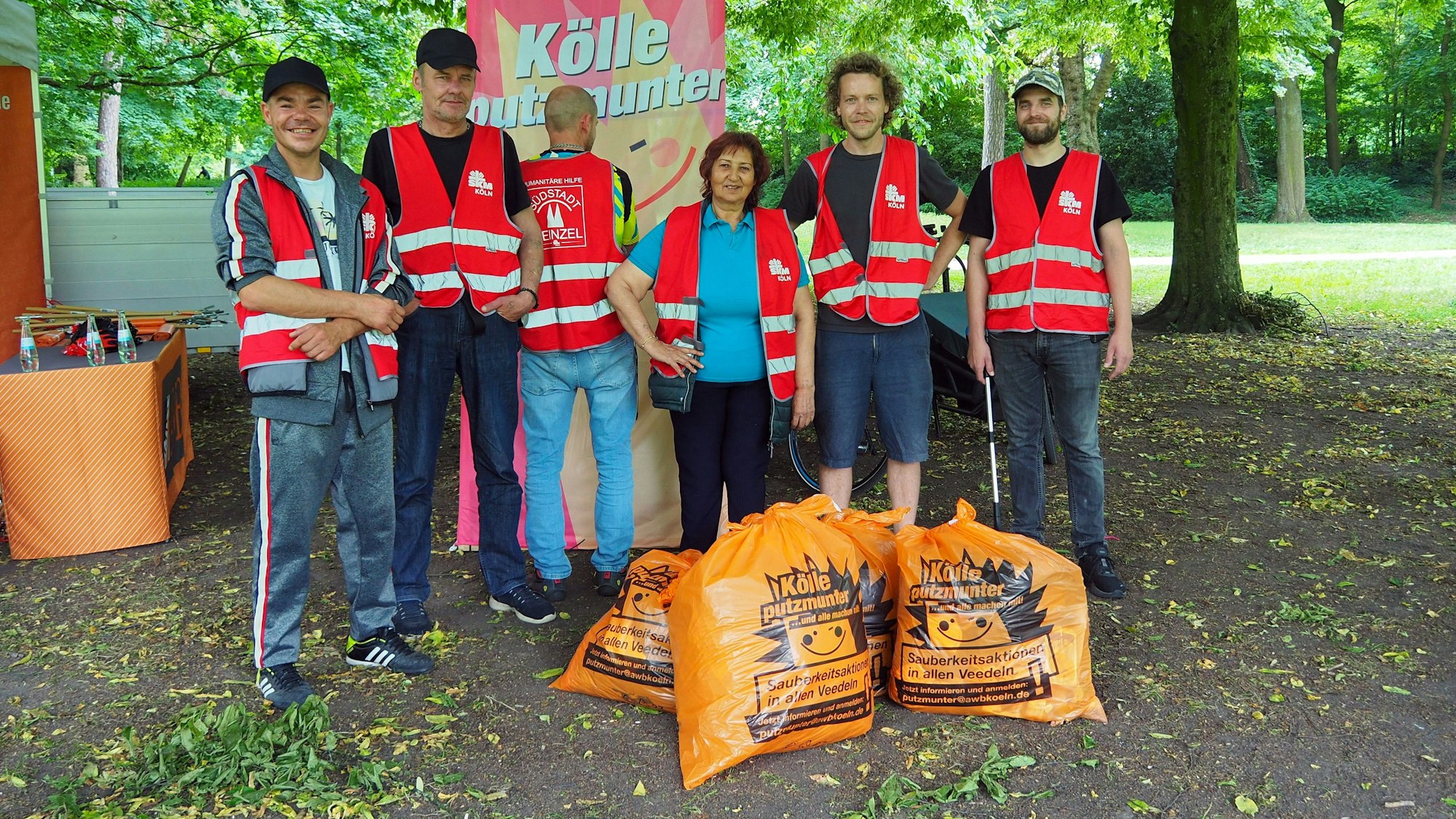 Sechs Menschen in rot-weißen Signalwesten stehen hinter einigen prallgefüllten Müllsäcken im Park.