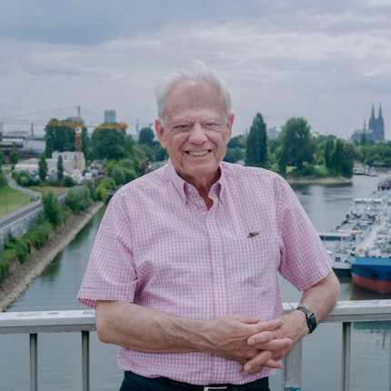 Ein Mann im rosa-weißen Karohemd steht auf einer Brücke. Im Hintergrund sind das Mülheimer Hafenbecken und der Dom zu sehen.